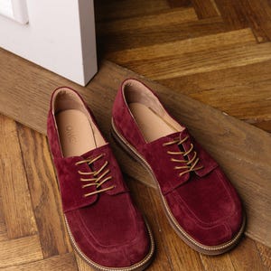 May include: A pair of deep burgundy suede lace-up shoes featuring a rounded toe box, tan laces, and a light-colored sole with visible stitching. The interior of the shoes is lined in a light tan material with the brand name OMG printed on the insole.