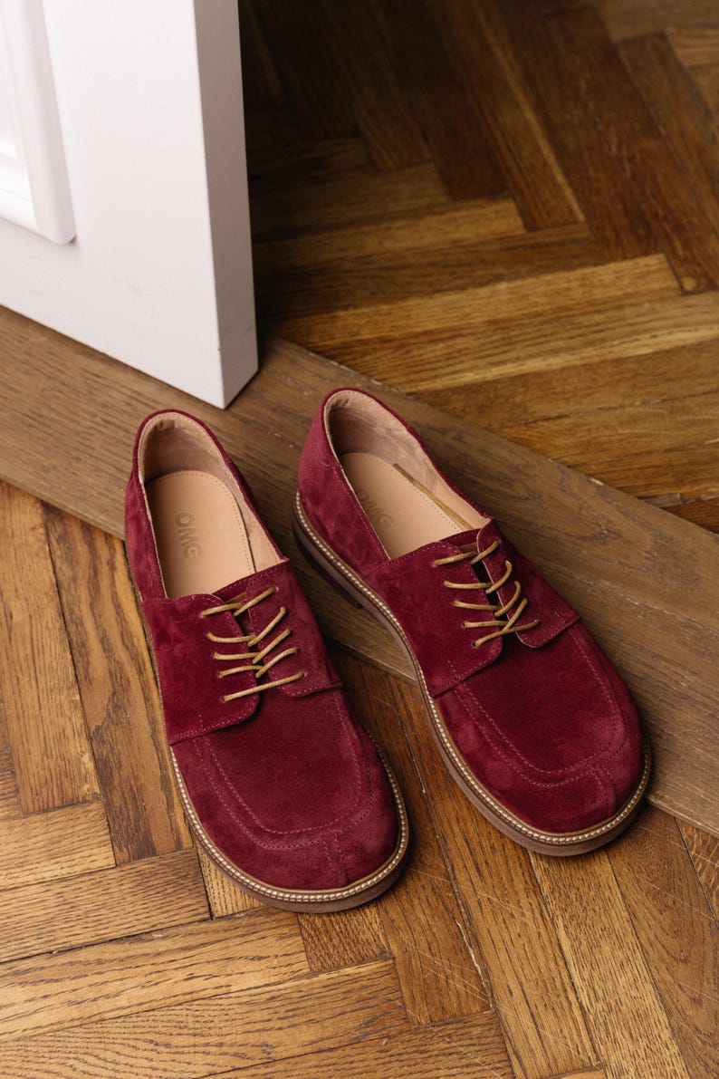 May include: A pair of deep burgundy suede lace-up shoes featuring a rounded toe box, tan laces, and a light-colored sole with visible stitching. The interior of the shoes is lined in a light tan material with the brand name OMG printed on the insole.