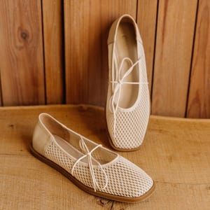 May include: A pair of beige ballet flats featuring a square toe, a mesh upper with a diamond-shaped pattern, and thin matching laces tied in a bow across the top. The shoes have a solid heel counter and a flat, brown rubber sole for stability.