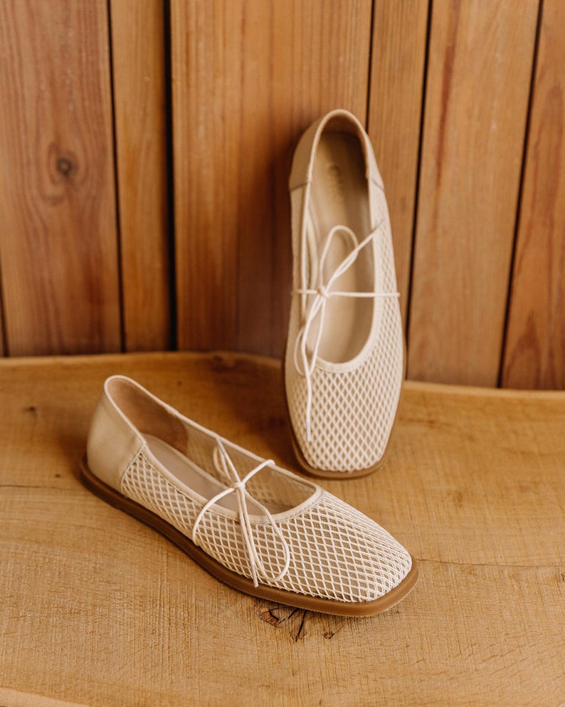 May include: A pair of beige ballet flats featuring a square toe, a mesh upper with a diamond-shaped pattern, and thin matching laces tied in a bow across the top. The shoes have a solid heel counter and a flat, brown rubber sole for stability.