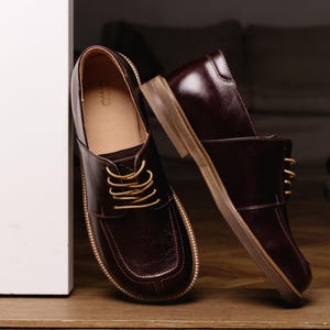 May include: A pair of dark burgundy leather shoes with a square toe design, featuring light tan laces and a tan sole. The interior of the shoe is lined in a light tan material with the brand name CWO visible on the heel of the inner sole.
