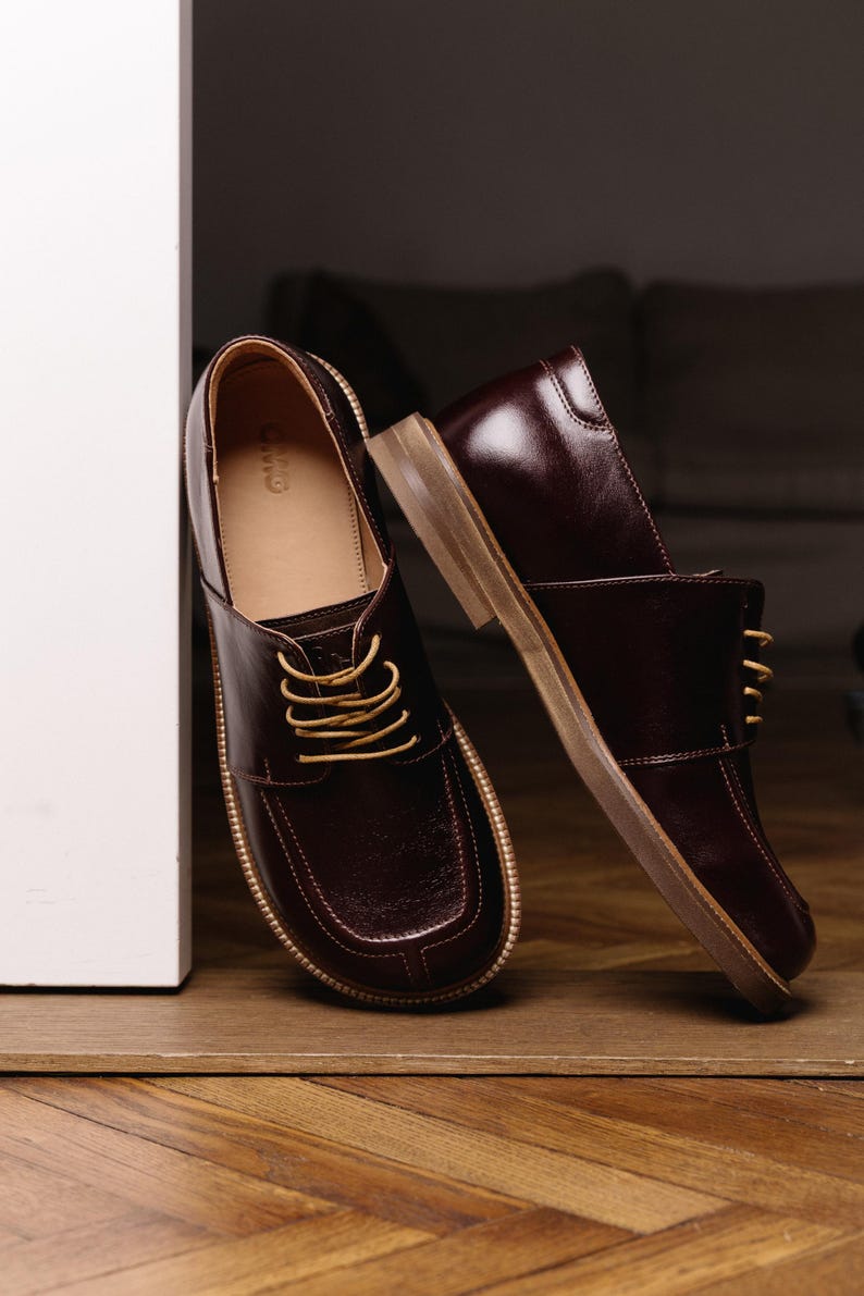 May include: A pair of dark burgundy leather shoes with a square toe design, featuring light tan laces and a tan sole. The interior of the shoe is lined in a light tan material with the brand name CWO visible on the heel of the inner sole.