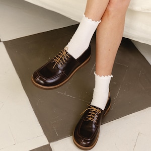 May include: A pair of dark brown leather oxford shoes with a rounded toe, a raised seam detail on the vamp, and thin tan laces. The shoes are worn with white ribbed crew socks that feature a delicate ruffled edge at the top of the cuff.