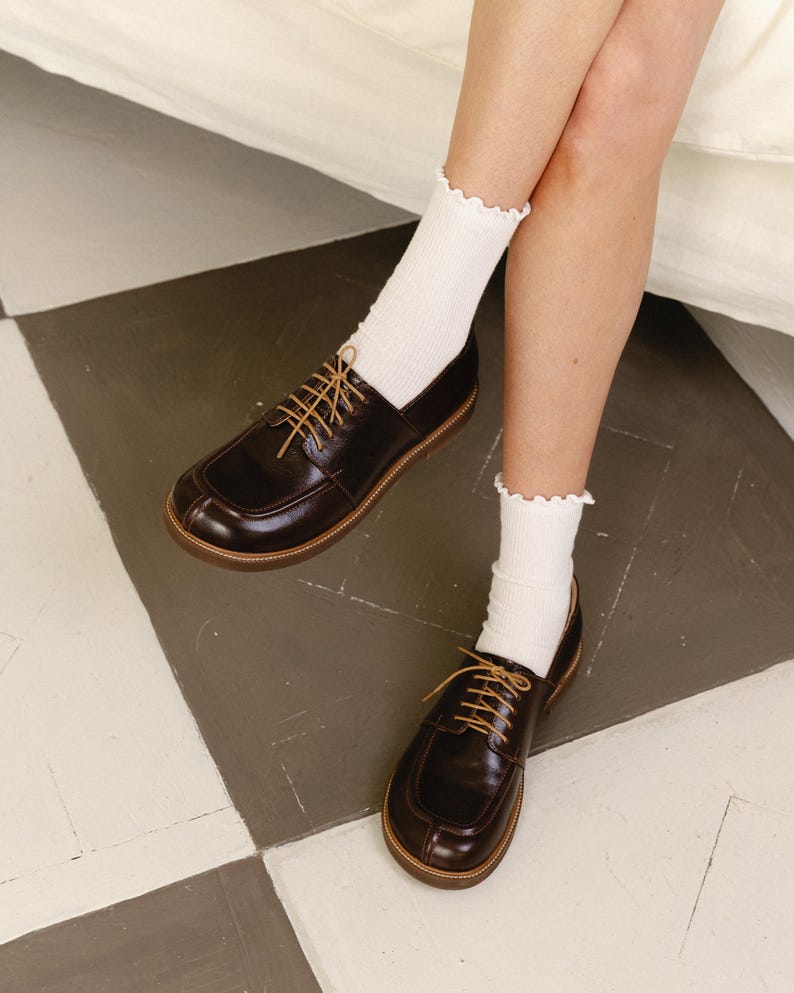 May include: A pair of dark brown leather oxford shoes with a rounded toe, a raised seam detail on the vamp, and thin tan laces. The shoes are worn with white ribbed crew socks that feature a delicate ruffled edge at the top of the cuff.