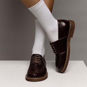May include: A pair of dark brown leather derby shoes with a moc-toe design and light tan laces, paired with white ribbed crew socks. The shoes feature a low heel and a visible welt construction, showcasing a polished finish and a classic silhouette.