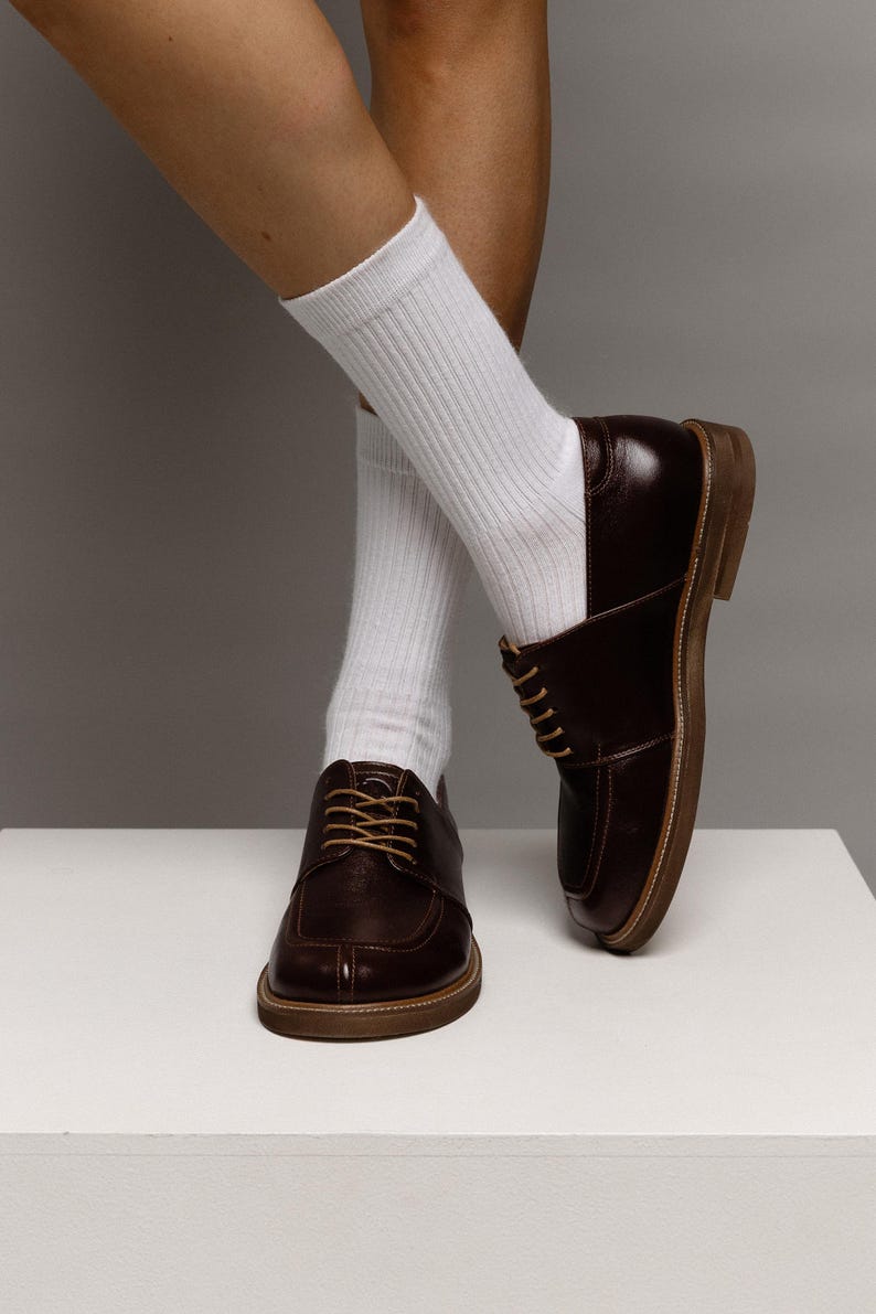 May include: A pair of dark brown leather derby shoes with a moc-toe design and light tan laces, paired with white ribbed crew socks. The shoes feature a low heel and a visible welt construction, showcasing a polished finish and a classic silhouette.