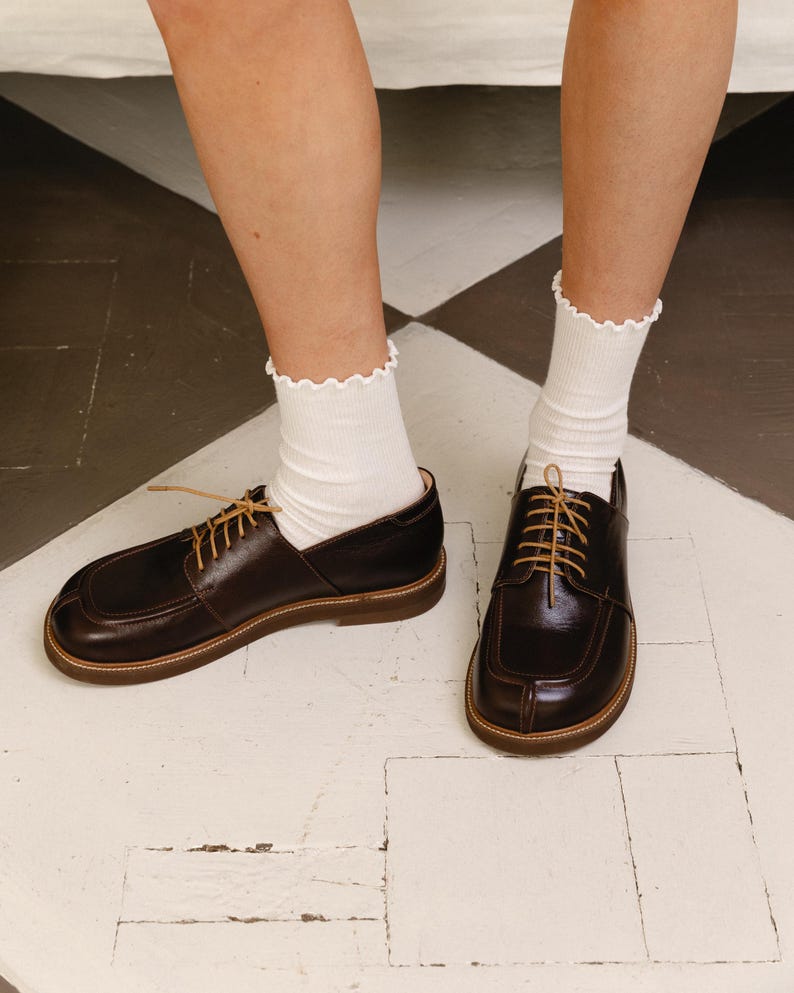 May include: A pair of dark brown leather oxford shoes with a square toe, visible stitching along the vamp, and thin tan laces. The shoes are paired with white ribbed crew socks featuring a delicate lettuce-edge trim at the top cuff.