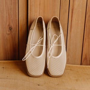 May include: A pair of cream-colored mesh ballet flats featuring a square toe, a thin lace-up tie detail across the top, and a flat brown sole. The interior of the shoe is lined in a matching cream tone with a visible brand logo on the heel bed.
