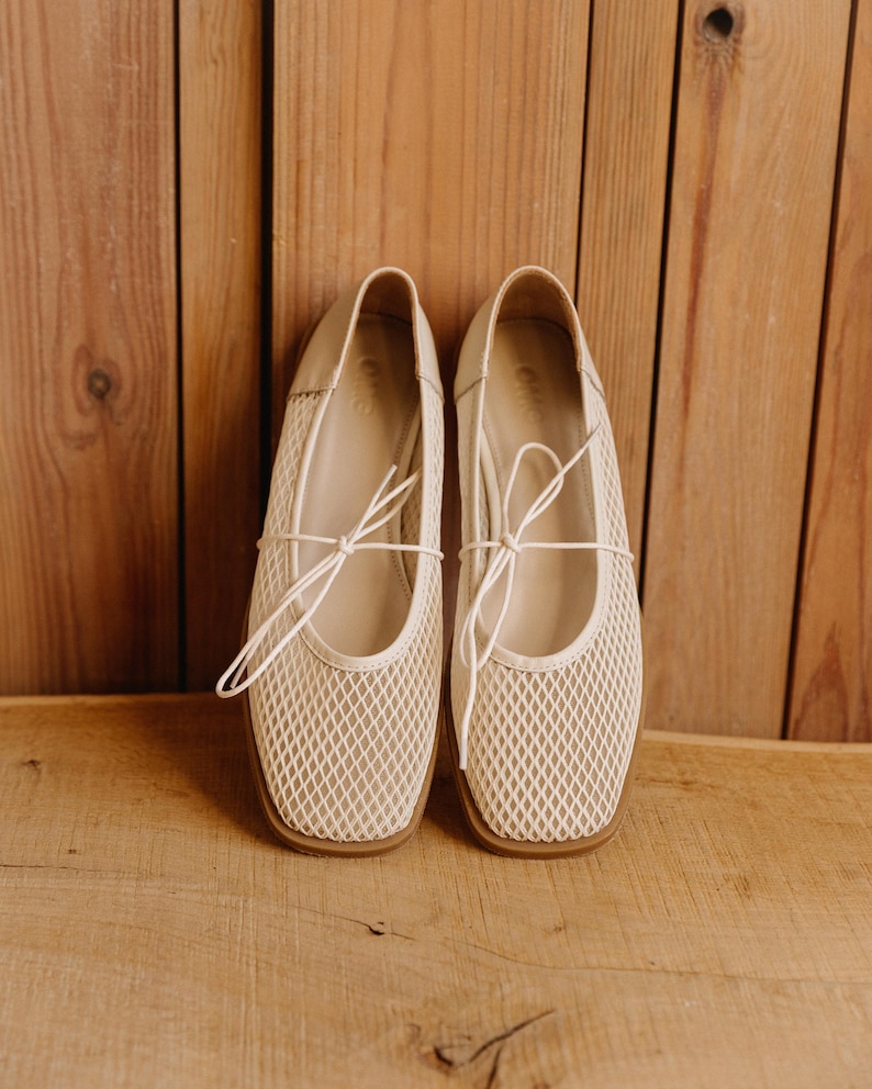 May include: A pair of cream-colored mesh ballet flats featuring a square toe, a thin lace-up tie detail across the top, and a flat brown sole. The interior of the shoe is lined in a matching cream tone with a visible brand logo on the heel bed.