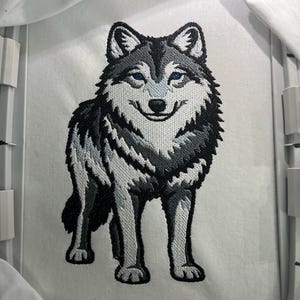 Pode incluir: Design de lobo bordado em preto, cinza e branco com olhos azuis. O lobo está em pé, voltado para a frente, e tem uma aparência detalhada e texturizada. O bordado está em um tecido branco, provavelmente para um patch ou aplicação.