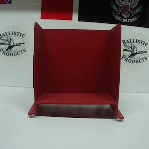 May include: A red metal shelf with a rectangular base and a back panel. The shelf has two side panels and is designed to hold small items. The background includes white surfaces with logos and flags.