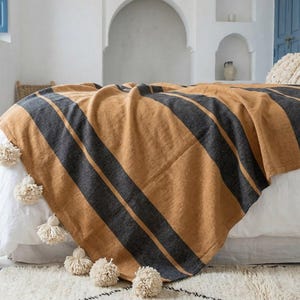 May include: A rectangular throw blanket featuring wide, alternating horizontal stripes in mustard yellow and charcoal grey. The blanket is finished with a series of cream-coloured decorative tassels attached along the bottom edge, adding texture to the fabric.