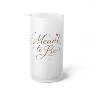 Puede incluir: Un vaso esmerilado blanco con las palabras "Meant to Be" en una fuente marrón. El vaso tiene forma cilíndrica y está decorado con pequeños detalles de estrellas y corazones.