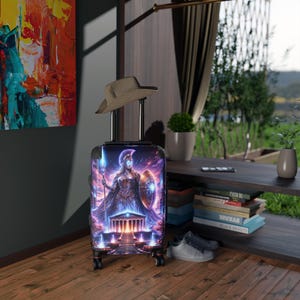 May include: A wheeled suitcase featuring a colourful illustration of a warrior goddess with a spear and shield, set against a temple and a cosmic sky. The suitcase has a retractable handle and four wheels. A straw hat sits on top.