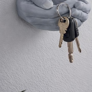 Op de afbeelding: Een grijze, aan de muur bevestigde handsculptuur die een set sleutels vasthoudt. De sleutels zitten aan een zilveren ring en hebben verschillende vormen en kleuren. De sculptuur is bevestigd aan een getextureerde witte muur, met een kleine potplant op de voorgrond.