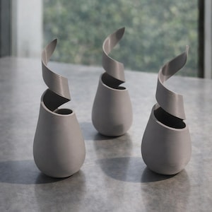 May include: Three gray, teardrop-shaped vases with a spiraling, ribbon-like design extending from the top. The vases are arranged on a gray surface, with a blurred green background visible through a window.