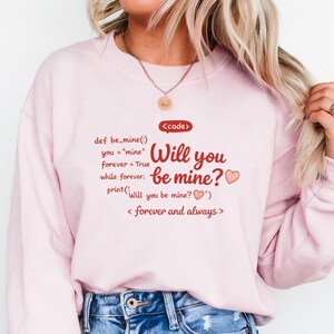 May include: A light pink sweatshirt with the text "Will you be mine?" in red, along with a heart graphic. The design includes code-like text and the phrase "forever and always."