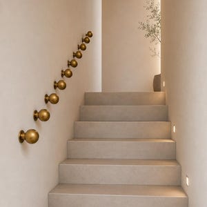 May include: A series of eight spherical brass wall-mounted handrail supports arranged in a diagonal line along a staircase. Each support features a round knob attached to a circular base plate, finished in a matte metallic gold tone.