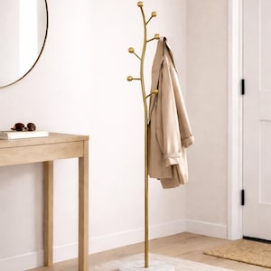 May include: A gold metal coat rack with a marble base. The rack has a tree-like design with multiple rounded knobs for hanging coats and other items. A tan coat hangs on the rack. The rack is in a room with a light-colored rug and a wooden table.