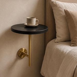May include: A wall-mounted floating side table featuring a circular black tabletop supported by a vertical brass-toned metal post and a circular wall bracket. A speckled beige ceramic mug sits on a matching saucer atop the black surface.