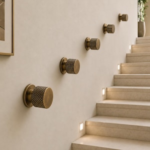 May include: A series of five cylindrical wall hooks made of brushed brass with a textured, diamond-patterned knurled finish. Each hook is mounted individually to the wall in a diagonal ascending line, featuring a flat circular face and base.