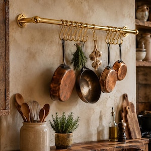 May include: A polished gold-toned metal wall-mounted rail featuring multiple matching S-shaped hooks. Hanging from the hooks are several copper-toned metal pans, a bundle of green herbs, and a cluster of garlic bulbs against a textured wall.