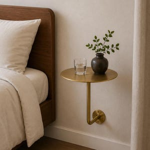 May include: A wall-mounted circular side table made of brushed brass with a single curved support arm. On the table surface sits a clear glass tumbler filled with water and a dark grey ceramic vase containing a small green leafy plant branch.