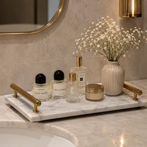 May include: A rectangular white marble tray with gold-toned ribbed metal handles. The tray holds three glass perfume bottles with black and gold caps, a small gold-lidded jar, and a ribbed ceramic vase containing white baby's breath flowers.