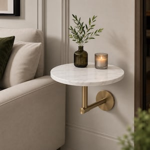 Puede incluir: Un estante flotante montado en la pared con una superficie circular de mármol blanco con vetas grises. El estante se sostiene mediante un soporte de metal dorado cepillado que se fija a la pared con una placa circular. Un jarrón y una vela están encima.