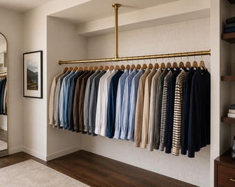 May include: A brass ceiling-mounted clothing rod holds a row of long-sleeved shirts and sweaters in shades of beige, blue, white, gray, and navy. The garments are arranged on wooden hangers against a white brick wall in a walk-in closet space.