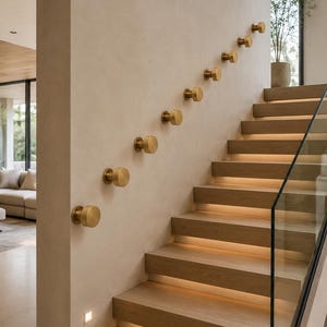 May include: A series of nine circular, brushed brass wall-mounted knobs arranged in a diagonal line along a beige wall, positioned adjacent to a wooden staircase with integrated step lighting and a glass railing panel.