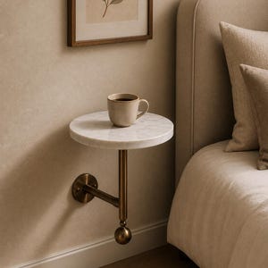 May include: A wall-mounted side table featuring a circular white marble top supported by a brass-toned metal arm and a spherical finial at the base. A beige ceramic mug filled with a dark liquid sits on the marble surface of the table.