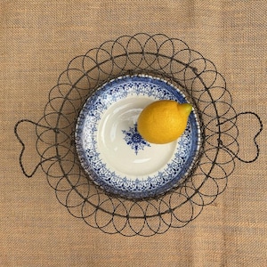 May include: A vintage-style serving tray with a decorative wire frame, holding a blue and white patterned plate. A bright yellow lemon rests on the plate. The tray sits on a burlap surface.