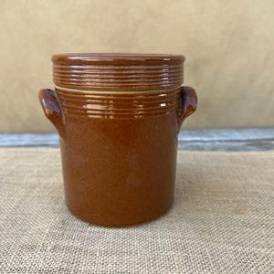 May include: A brown ceramic jar with a lid and two small handles. The jar has a textured surface and a cylindrical shape. The lid has horizontal ridges. The jar is likely used for storage or decoration.