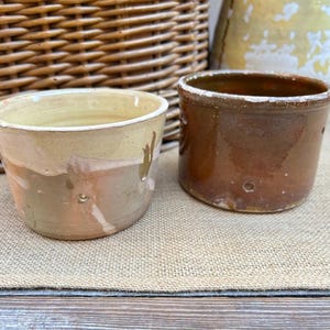 May include: Two small, rustic ceramic pots. One is light tan with a brown and peach band, the other is a dark brown with a glossy finish. Both have a distressed, aged appearance, suggesting they are vintage or antique.