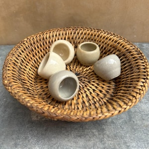 May include: A round, woven brown wicker basket containing five small, smooth, spherical ceramic vessels in varying shades of off-white, cream, and light gray. Each ceramic piece features a wide, open circular top and a rounded, bulbous base.