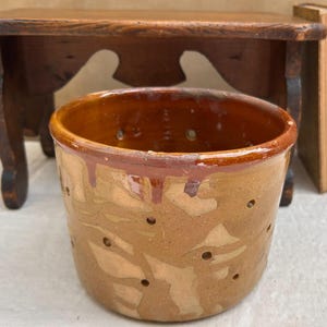 May include: A handmade, rustic ceramic pot with a brown and tan glaze. The pot has a series of small holes around the sides and a dark brown rim. The pot is sitting in front of a small wooden table and some old books.