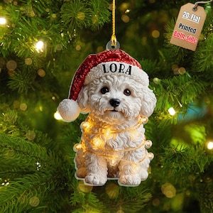 May include: A white curly-haired dog ornament wearing a red Santa hat with the name LORA printed on the white brim. The dog is wrapped in a string of small glowing warm white fairy lights. A brown tag at the top reads 2D-Flat Printed Both-side.