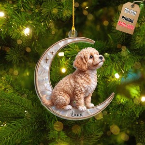 Op de afbeelding: Een ornament in de vorm van een halve maan met een lichtbruine krulharige puppy die op de boog zit. De maan heeft een parelmoer afwerking met kleine steraccenten en de naam Luna in zwarte letters onderaan de halve maan.