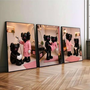 Op de afbeelding: Drie ingelijste wandkunstprints met zwarte cartoonfiguren met X-vormige ogen. De figuren zijn afgebeeld in een badkamer, dragen roze badjassen, houden een douchekop vast, dragen een ander figuur of zitten op een toilet.