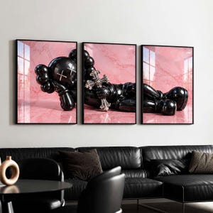 May include: A triptych wall art set featuring a glossy black character with X-shaped eyes lying down against a pink marble background. The central panel shows the figure holding a silver cross. Each piece is framed in a thin black border.