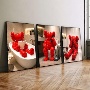 Könnte beinhalten: Ein Set aus drei gerahmten Wandbildern, die eine glänzend rote Zeichentrickfigur in verschiedenen Badezimmerszenen zeigen. Ein Bild zeigt die Figur in einer Badewanne beim Lesen, ein anderes auf einer Toilette mit einem Telefon und ein drittes am Boden.