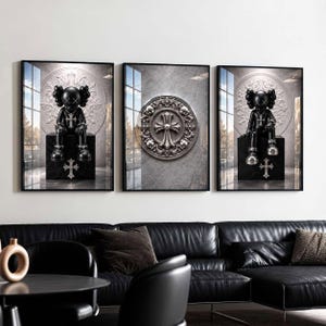May include: A set of three framed wall art prints. The outer two prints feature a black cartoon character with X-shaped eyes sitting on a black pedestal with a silver cross emblem. The center print displays a circular silver cross medallion.