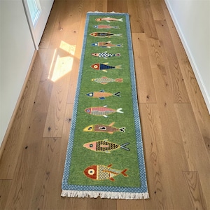 May include: A green runner rug with a blue border and white fringe. The rug features a repeating pattern of colorful fish in various designs. The rug is placed on a wooden floor.