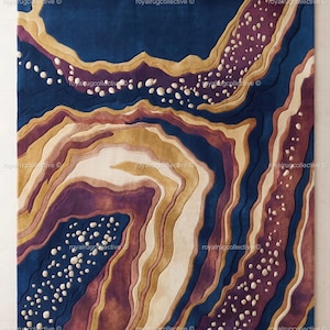 Può includere: Un tappeto rettangolare caratterizzato da un design astratto a strati con forme fluide e organiche in blu navy profondo, viola prugna intenso, beige caldo e crema. La superficie presenta accenti testurizzati in rilievo color bianco.