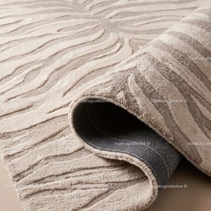 May include: A beige and cream area rug featuring a raised, textured, wavy zebra-like pattern. The rug is partially folded over, revealing a dark gray, textured backing material. The surface has a soft, plush pile with repeating organic lines.