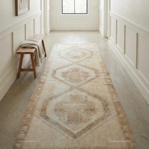 May include: A long, rectangular rug with a geometric pattern in beige and brown tones. The rug is placed in a hallway with white walls, a wooden bench, and a window. The rug's design features repeating diamond and octagon shapes.