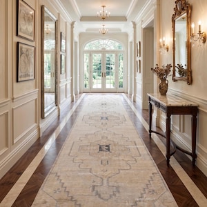 May include: A long, rectangular area rug with a beige base and faded blue geometric patterns, featuring a central medallion and fringed edges, laid out on a dark wood floor in a bright, high-ceilinged hallway with white paneled walls.