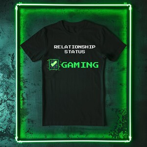 May include: Black t-shirt with the text "RELATIONSHIP STATUS" above "GAMING" in pixelated green font. A green checkmark graphic is next to the word "GAMING". The shirt is displayed against a dark green background with a neon green border.