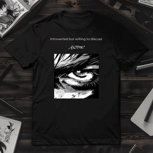 以下が含まれることがあります： 黒いTシャツ。白黒の目のイラストが特徴です。「Introverted but willing to discuss ANIME」の文字が目のグラフィックの上にプリントされています。柔らかい素材で作られたシャツです。